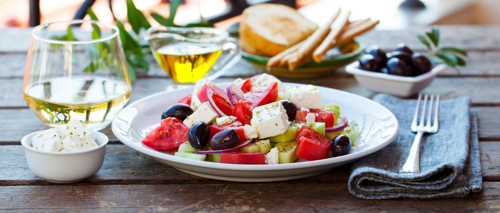 Plato vegano de ensalada griega con verduras frescas, queso, aceitunas negras y vino blanco al fondo.